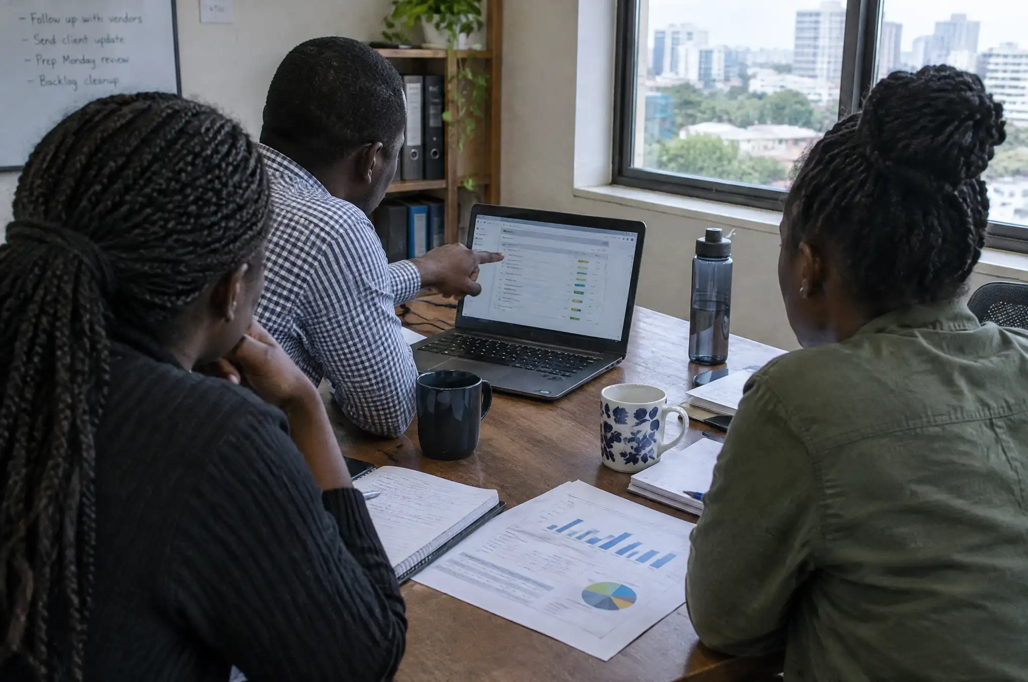 Three people at a meeting table look at a task list on a laptop, with printed charts and notebooks in front of them
