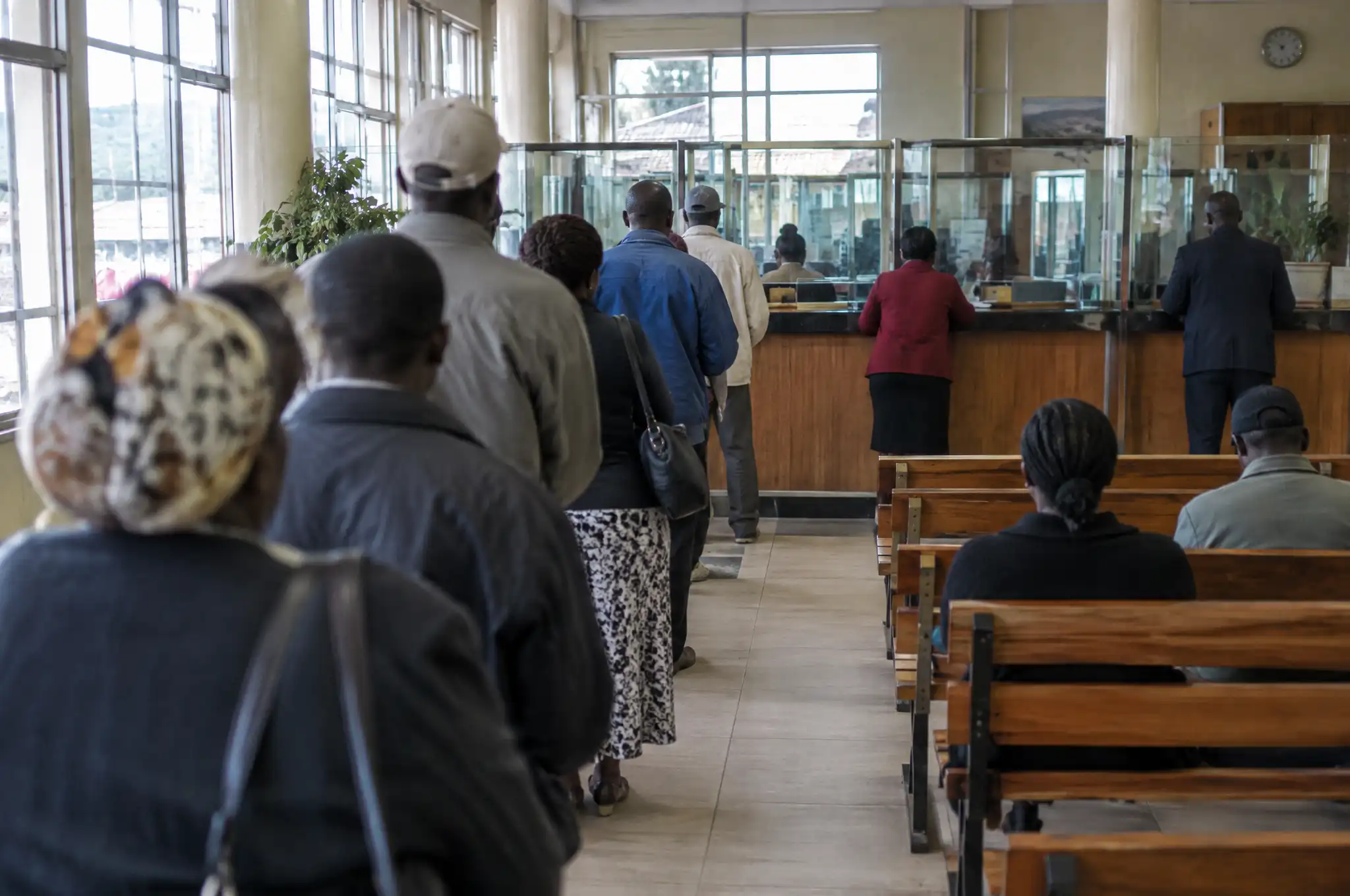 People queue at the counters of a banking hall while others wait on wooden benches